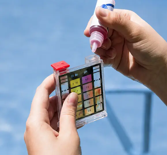 Hands holding a swimming pool test kit and adding reagent drops to the water sample chamber.