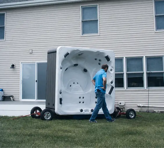 A man pushes a hot tub on a flatbed dolly across a lawn toward a house.
