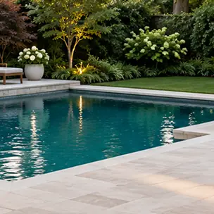 A rectangular swimming pool surrounded by a stone patio and a garden with flowering shrubs.