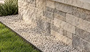 A stone retaining wall sits above a garden bed filled with light-colored pebbles and bordered by a strip of green grass.