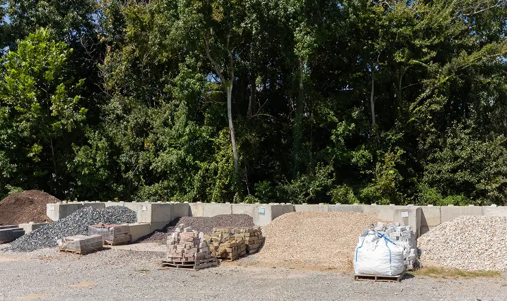 Piles of various landscaping materials, including gravel, stone pavers, and sand, organized in concrete bays in front of a line of trees.