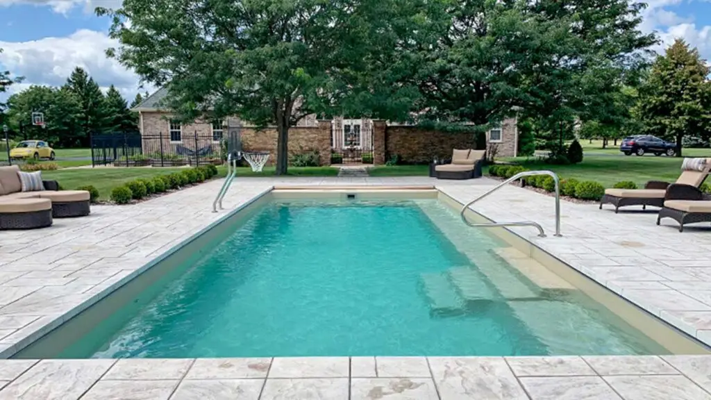 Rectangular swimming pool with a stone patio, pool furniture, and a basketball hoop in a residential backyard.
