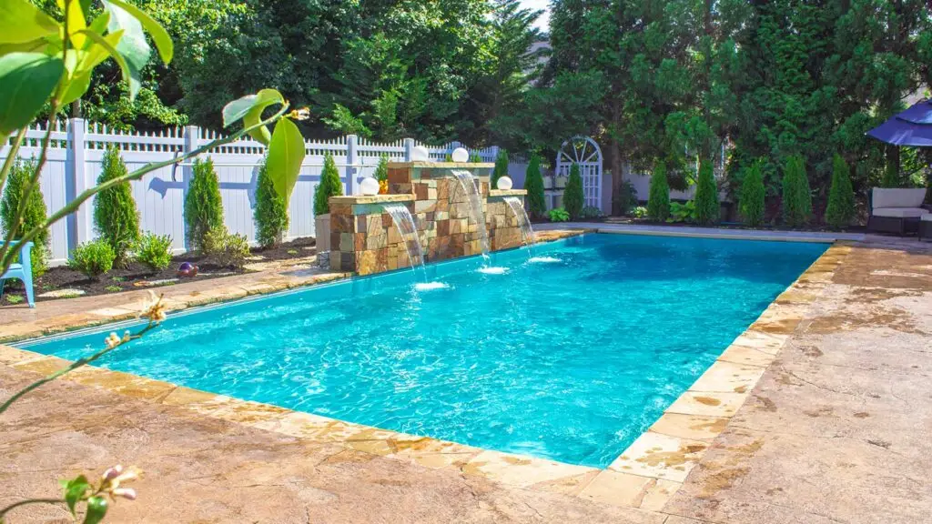 A rectangular swimming pool with a stone water feature and a white fence in a backyard.