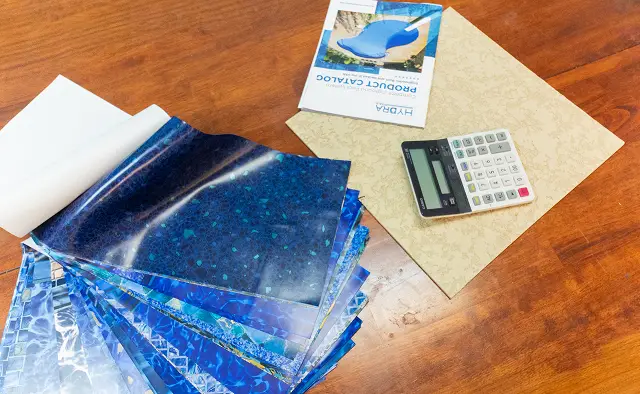 A fan of blue swimming pool liner samples, a product catalog, and a calculator resting on a square tile sample.