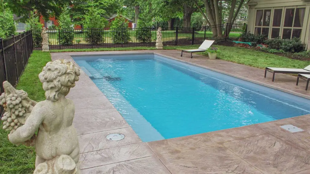 A rectangular swimming pool surrounded by a stone patio, lawn, and decorative stone statues in a fenced backyard.