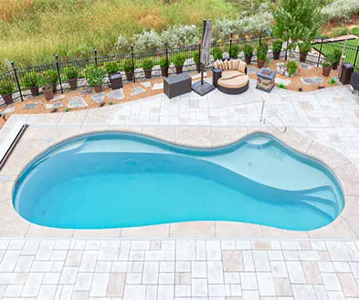 An elevated view of a kidney-shaped swimming pool with built-in stairs, set in a stone patio with outdoor seating and a fenced garden border.