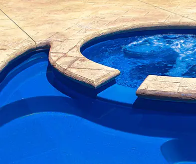A blue pool with a connected, bubbling hot tub surrounded by stamped concrete decking.