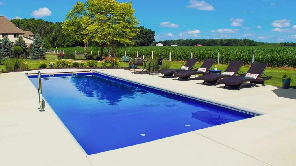 A rectangular backyard swimming pool surrounded by a concrete patio with four lounge chairs and a small dining set, situated next to a large cornfield.