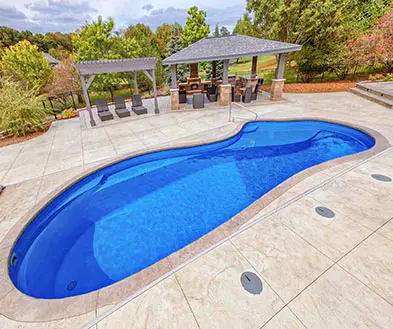 A kidney-shaped swimming pool surrounded by a concrete patio, with an outdoor pavilion and dining area in the background.