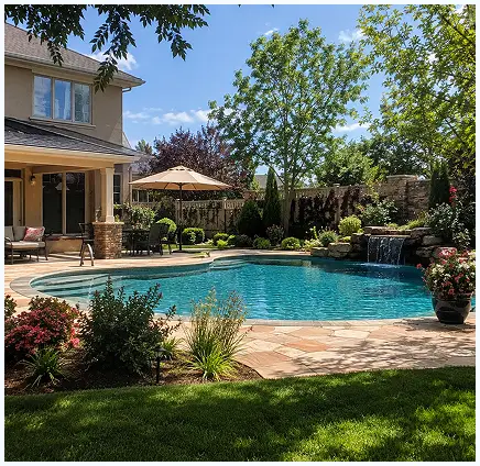 A backyard swimming pool with a stone waterfall feature, surrounded by a paved patio, landscaping, and a house with a covered porch.
