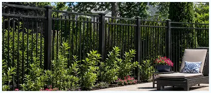 A black metal fence stands behind a row of green shrubs and a patio chair with a cushion and a potted plant.