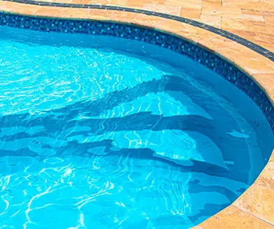 A curved swimming pool with blue water and built-in entry steps, bordered by stone decking.