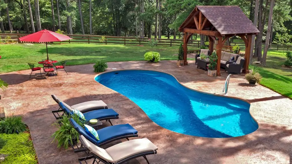 A kidney-shaped swimming pool with a stone patio, lounge chairs, a dining set under a red umbrella, and a wooden gazebo.