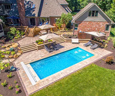 Aerial view of a rectangular backyard swimming pool surrounded by a stone patio, outdoor seating, and a brick house.