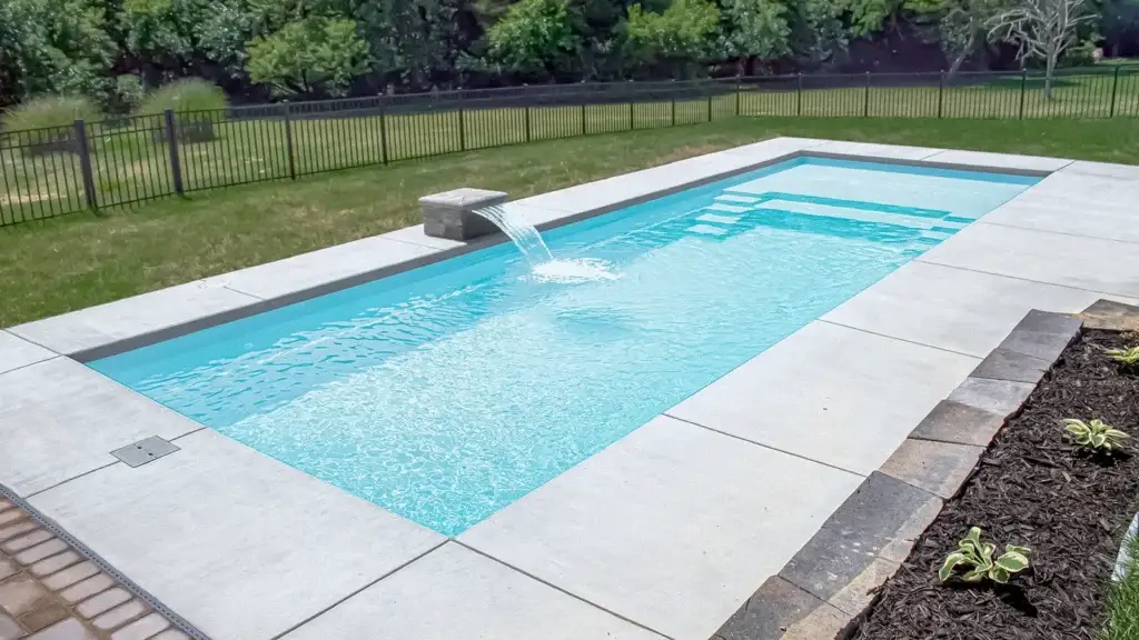 A rectangular swimming pool with a stone water feature, surrounded by a concrete deck and a black metal fence in a grassy yard.