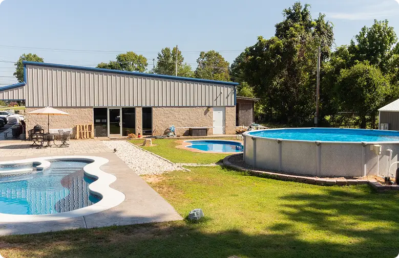 Three outdoor swimming pools in a grassy backyard next to a large metal building.