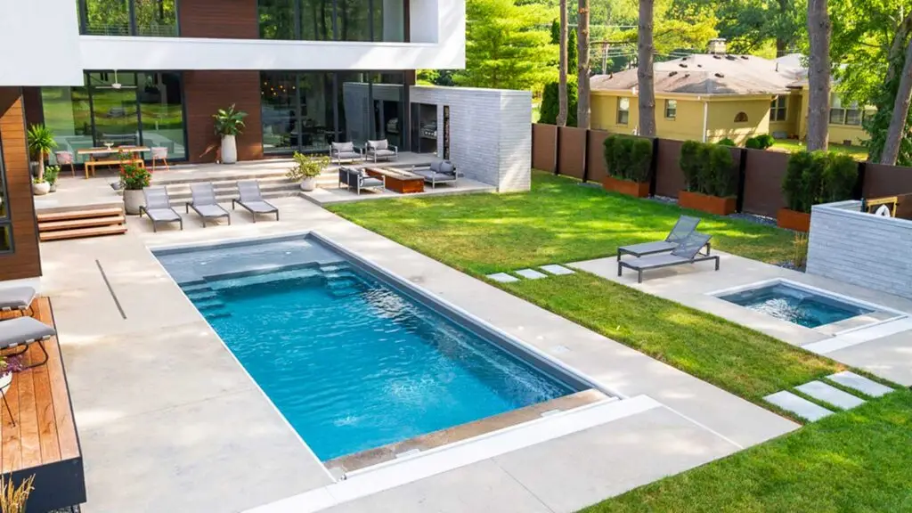 A modern backyard featuring a rectangular swimming pool, a separate hot tub, and several seating areas on a concrete patio next to a multi-story house.