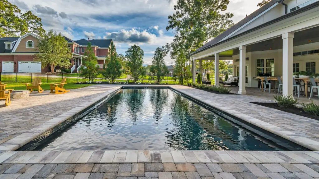 A rectangular swimming pool surrounded by a stone patio and a covered outdoor seating area.