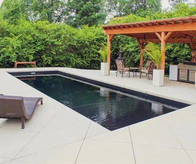 A rectangular swimming pool with a dark interior set into a light-colored patio next to a wooden pergola with outdoor seating.